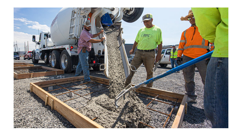 Construction workers pour concrete from a large mixing truck into a frame on the ground.