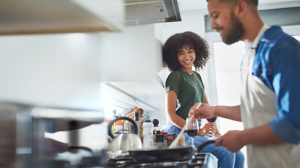 Smiling woman sitting on kitchen counter with a glass of wine watches man cook food